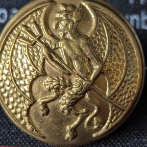 May include: A round, antique brass button with a detailed relief design. The central image features a winged, demonic figure holding a torch, with a lion-like creature below. The button has a textured edge and a slightly worn appearance.