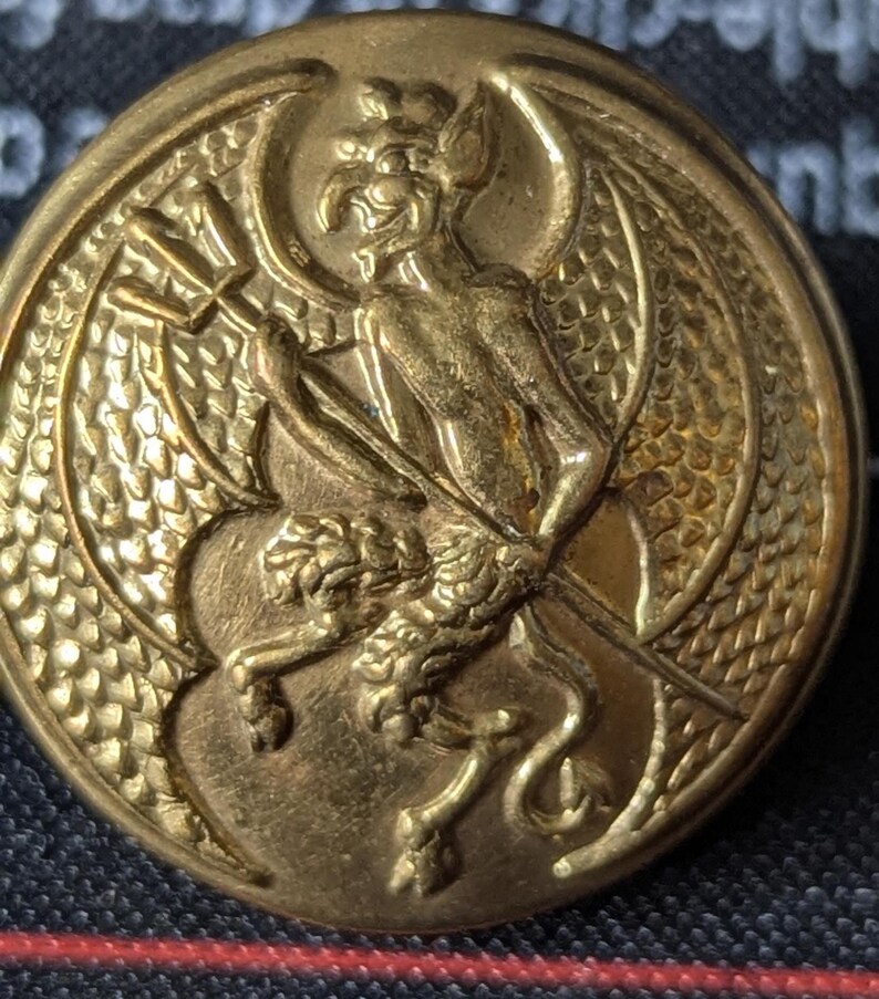 May include: A round, antique brass button with a detailed relief design. The central image features a winged, demonic figure holding a torch, with a lion-like creature below. The button has a textured edge and a slightly worn appearance.