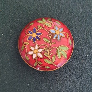 Op de afbeelding: Een ronde, rode emaille-speld met een bloemenmotief. De bloemen zijn blauw, wit en groen.