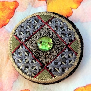 May include: A round, embroidered brooch with a central green button. The design features a geometric pattern in gray, red, and olive green thread on a black base. The brooch has a textured, handcrafted appearance.