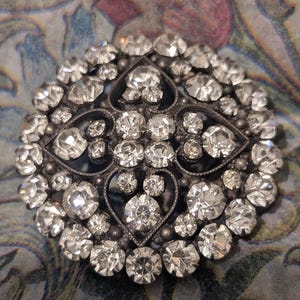 May include: Circular, antique-style brooch with numerous clear, faceted rhinestones set in a dark metal frame. The brooch features a central design with heart-shaped elements and a detailed, ornate border. A vintage jewelry piece.