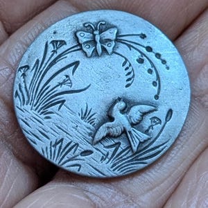 Op de afbeelding: Een ronde, zilverkleurige metalen knoop met een reliëf natuurtoneel. Het ontwerp toont een vlinder, een vogel in vlucht en gestileerde planten. De knoop wordt in een hand gehouden, wat de grootte en textuur laat zien.