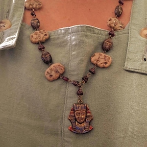 May include: A beaded necklace featuring a pendant of an Egyptian pharaoh. The necklace has tan, brown, and red beads, with a gold-colored chain. The pharaoh pendant is detailed with blue, red, and gold accents.