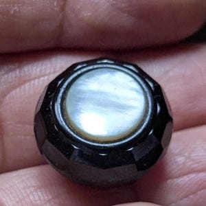 May include: A black, faceted button with a round, iridescent mother-of-pearl centre. The button has a geometric design and a polished finish, suitable for sewing or crafts.