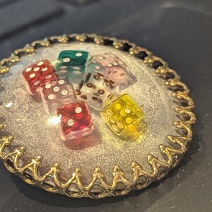 May include: A round, gold-toned pendant with a clear resin center, showcasing six colorful dice. The dice are red, green, yellow, and clear with white dots. The pendant has a decorative, ornate edge.