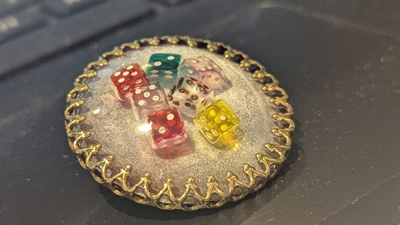 May include: A round, gold-toned pendant with a clear resin center, showcasing six colorful dice. The dice are red, green, yellow, and clear with white dots. The pendant has a decorative, ornate edge.