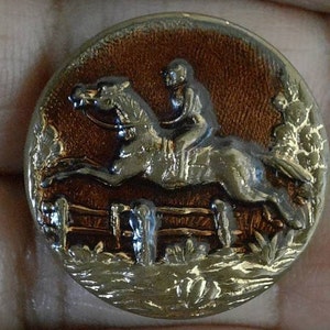 May include: A gold-toned metal button with a raised design of a horse jumping over a fence. The horse is brown and gold, and the rider is wearing a brown and gold uniform. The background is a brown and gold color.