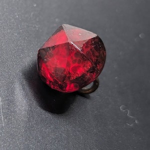 May include: A faceted, deep red gemstone with a metallic loop. The gem has a complex, internal structure, and a brilliant, reflective surface. The gem is set against a dark background.
