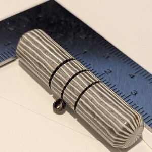 May include: A cylindrical, striped object with a metal loop. The object is primarily gray with white stripes and has three segmented rings. A ruler is in the background, with measurements in inches.