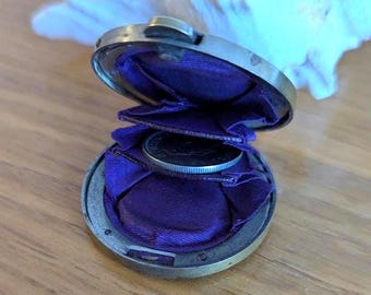 May include: A round, metallic coin purse with a hinged lid, featuring a deep purple fabric interior lining. The open container reveals a single silver-colored coin resting within the pleated purple fabric compartments, showcasing a compact design.
