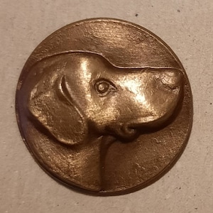 May include: A bronze-coloured metal coin featuring a detailed relief sculpture of a dog's head. The dog is facing to the right and has a prominent nose and ears.