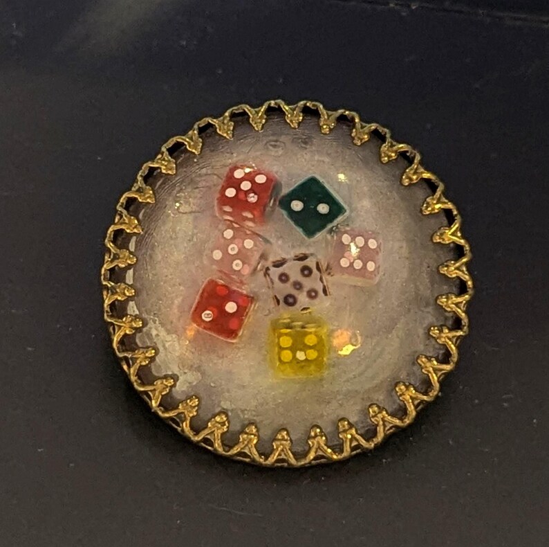 May include: A round, gold-colored brooch with a scalloped edge. Inside, a clear resin holds several colorful dice. The dice are red, pink, green, and yellow, each with white dots. A unique, handcrafted accessory.
