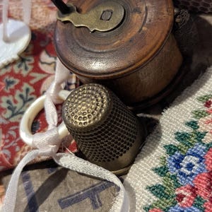 May include: A dark brown wooden spool with a brass winding handle, a metallic thimble with a textured surface, white ribbon, and a piece of fabric featuring a floral pattern in blue, pink, and green, alongside a small white box with text.