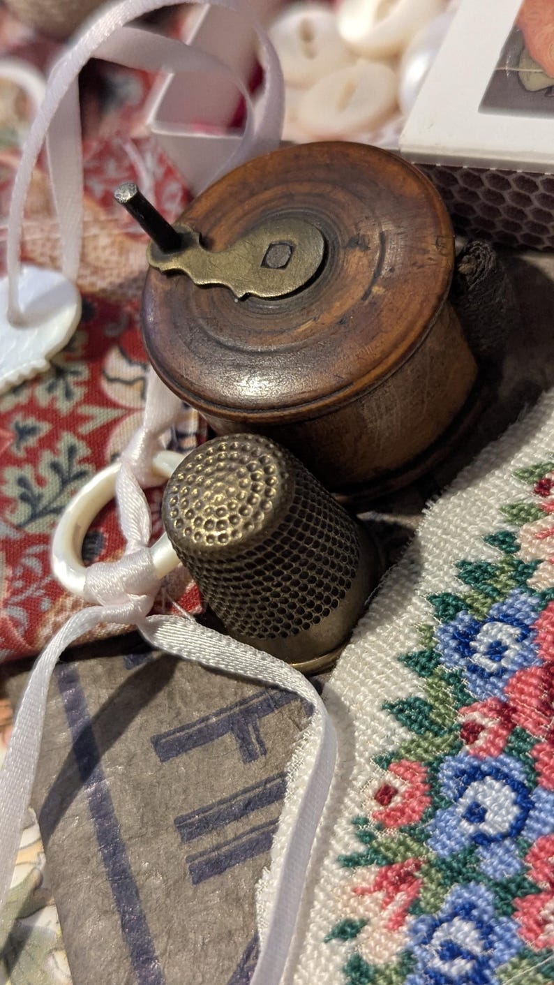 May include: A dark brown wooden spool with a brass winding handle, a metallic thimble with a textured surface, white ribbon, and a piece of fabric featuring a floral pattern in blue, pink, and green, alongside a small white box with text.