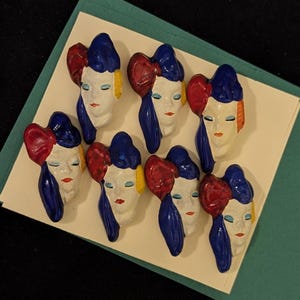 May include: Eight decorative face-shaped pins with a bow, painted in red, white, blue, and yellow. The pins are arranged on a white card, set against a dark background. The faces have closed eyes and red lips.