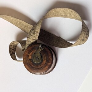 May include: A vintage circular wooden tape measure with a dark brown finish and a central metal winding crank. A long, beige fabric measuring tape with visible black markings extends from the spool, showing imperial units in inches.