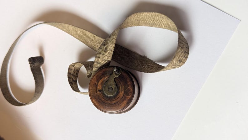 May include: A vintage circular wooden tape measure with a dark brown finish and a central metal winding crank. A long, beige fabric measuring tape with visible black markings extends from the spool, showing imperial units in inches.