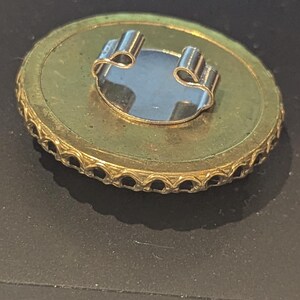 May include: A vintage gold-toned brooch with a circular design. The brooch features a decorative edge and a silver-colored clasp on the back. The center has a reflective, oval-shaped element. The brooch is set against a dark background.
