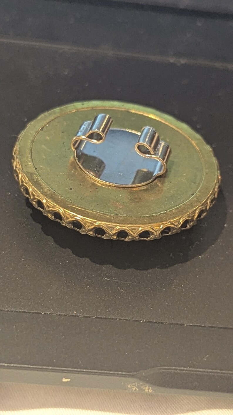 May include: A vintage gold-toned brooch with a circular design. The brooch features a decorative edge and a silver-colored clasp on the back. The center has a reflective, oval-shaped element. The brooch is set against a dark background.
