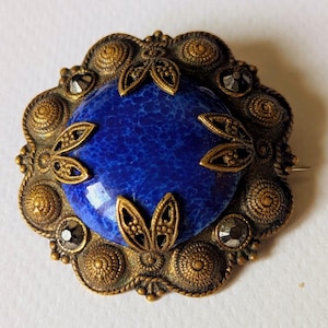 May include: Vintage brooch with a central blue stone, surrounded by an ornate, gold-toned metal frame. The frame features detailed circular and leaf-like designs, with small, dark accents. A pin is attached to the back.