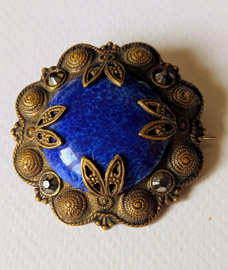May include: Vintage brooch with a central blue stone, surrounded by an ornate, gold-toned metal frame. The frame features detailed circular and leaf-like designs, with small, dark accents. A pin is attached to the back.