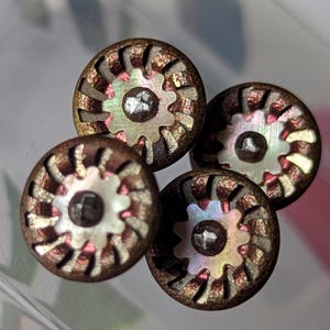 Puede incluir: Primer plano de cuatro botones decorativos. Cada botón tiene un anillo exterior de color bronce con un diseño en forma de engranaje y una forma central de engranaje iridiscente y nacarado. Los botones son probablemente para coser o manualidades.