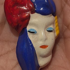 May include: A small, hand-painted ceramic brooch shaped like a human face. The face is cream-colored with light blue eyeshadow, closed eyes, and orange lips. It features a large, textured red and blue headpiece and a yellow accent on the side.