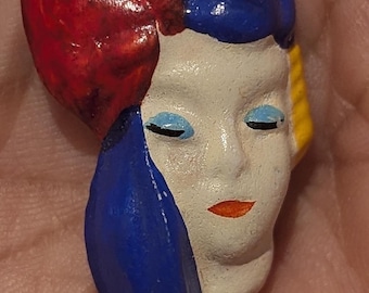 May include: A small, hand-painted ceramic brooch shaped like a human face. The face is cream-colored with light blue eyeshadow, closed eyes, and orange lips. It features a large, textured red and blue headpiece and a yellow accent on the side.