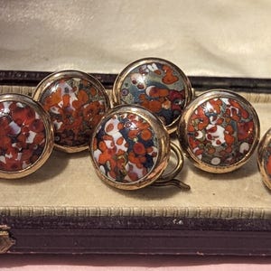 Op de afbeelding: Een set van vijf ronde knopen met een veelkleurig gespikkeld patroon van oranje, wit en donkerblauw, gevat in een goudkleurige metalen rand. Elke knoop heeft een glad, glanzend oppervlak en een cirkelvormig ontwerp met een abstract patroon.