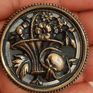 Puede incluir: Un botón vintage con un diseño negro y dorado que presenta una cesta de flores y un sombrero. El botón tiene una superficie texturizada en relieve y un borde de color plateado.