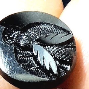 Puede incluir: Un botón negro con un diseño blanco de un pájaro estilizado con las alas extendidas. El botón tiene una superficie texturizada.