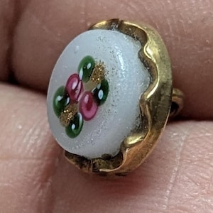 Può includere: Un piccolo bottone rotondo con una superficie bianca con un motivo floreale in verde, rosa e oro. Il bottone è incorniciato da un bordo dorato smerlato. Il bottone è probabilmente per cucire o fare artigianato.