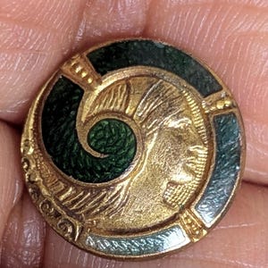 May include: A gold-toned, circular button with a green enamel border and a central design featuring a profile of a person. The button has a detailed, vintage aesthetic, with intricate patterns and a textured surface.
