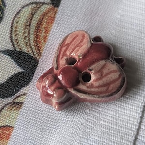 May include: A ceramic button shaped like a butterfly with pink and burgundy accents. The button has two holes for sewing.