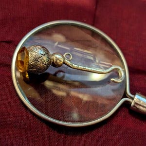 Puede incluir: Un alfiler de kilt de metal plateado con una cabeza texturizada en forma de cardo, rematada con una piedra facetada de color ámbar. El alfiler tiene un cierre de gancho curvado y muestra pequeños y claros sellos grabados en el eje.