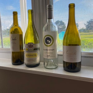 May include: Four wine bottles on a windowsill. From left to right: a red wine bottle labeled "Dante Cabernet Sauvignon", a white wine bottle labeled "Rombaur Carasvan Chardonnay", a white wine bottle labeled "Slingshot Napa Valley Sauvignon Blanc", and a white wine bottle labeled "Amarella Napa Valley Chardonnay".