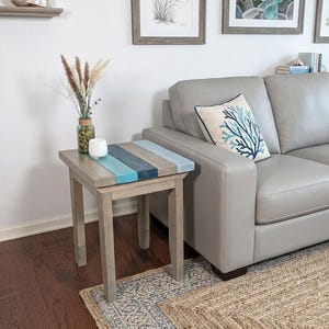 May include: A wooden side table with a gray finish and a top featuring painted blue and gray stripes. The table is square with four legs and is placed next to a gray sofa. A decorative pillow with a blue coral design rests on the sofa.