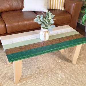 May include: A rectangular coffee table with a multi-colored top and light-colored legs. The top features horizontal stripes of various colors, including green, brown, white, and beige. A white vase with greenery sits on the table.