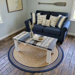 May include: A rectangular coffee table with a top made of weathered, multi-colored wooden planks and four solid white legs. The table is positioned on a round, natural-colored jute rug featuring concentric dark blue circular stripes.