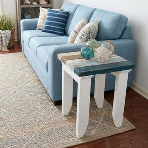 May include: A coastal-themed living room scene featuring a light blue sofa with decorative pillows. A small, square side table with a painted wood top in shades of blue, white, and tan sits in front of the sofa. Decorative glass balls and a starfish accent the table.