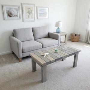 May include: A coastal-themed living room with a gray sofa, a wooden coffee table, and framed artwork. The coffee table features a starfish and decorative items. The room has a neutral color palette with a light gray carpet and white walls.