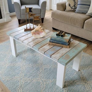 May include: A rectangular coffee table with a slatted top featuring a distressed finish in shades of blue, white, and natural wood. The table has four solid white legs and is decorated with a large seashell, a metal flag ornament, and books.