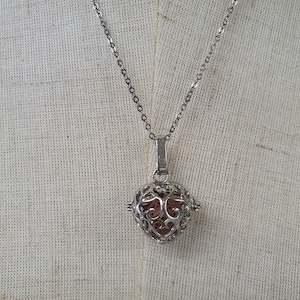 Pode incluir: Um colar de corrente de prata com um pingente em forma de gaiola de coração. A gaiola é feita de metal delicado e tem uma pequena pedra redonda rosa dentro.