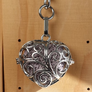 Può includere: Un ciondolo a forma di cuore in argento con un delicato design a filigrana. Il ciondolo è attaccato a una catena d'argento e pende da una superficie in legno.