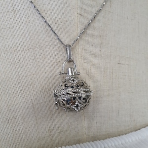 Könnte beinhalten: Eine silberfarbene Halskette mit einem dekorativen Anhänger. Der Anhänger ist als kugelförmiges Gitterdesign gestaltet und mit kleinen, klaren Kristallen besetzt. Die Halskette hängt an einer dünnen, silbernen Kette.