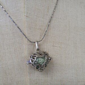May include: A silver heart-shaped locket necklace with a green lava rock inside. The locket has a delicate filigree design and a small latch on the side.