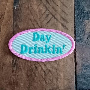 Puede incluir: Un parche bordado con las palabras "Day Drinkin'" en letras turquesas sobre un fondo azul claro. El parche tiene un borde rosa y una forma ovalada. El parche está sobre una superficie de madera.