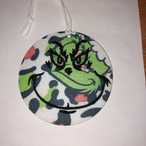 May include: A round, white, green, and black felt ornament with a green Grinch face embroidered on it. The ornament has a white string attached to it.