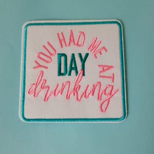 Puede incluir: Parche cuadrado blanco con borde turquesa y la frase "YOU HAD ME AT DRINKING" bordada en hilo rosa y turquesa. El parche está sobre un fondo azul claro.