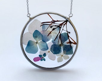 May include: A silver-toned necklace featuring a circular pendant. The pendant is a clear glass or resin circle, showcasing pressed blue, white, and pink flowers and foliage. The chain is delicate and hangs from the top of the pendant.